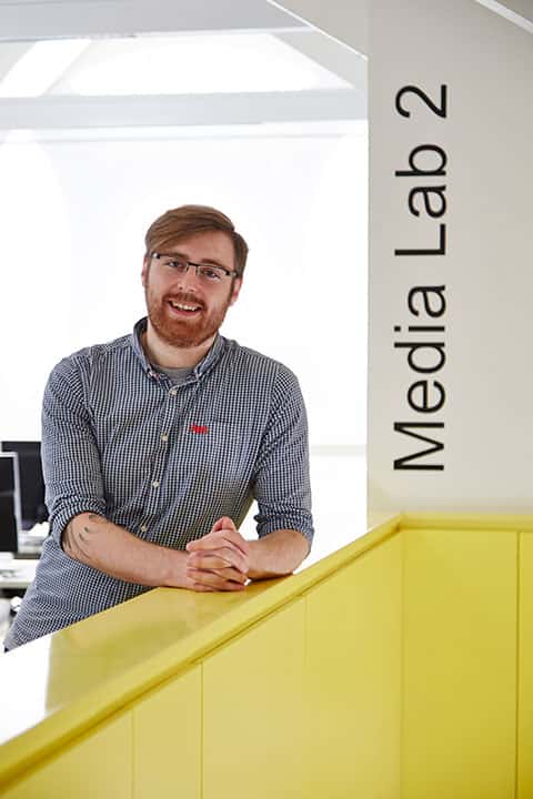 portrait photo of lecturer george beard with hands crossed and leaning on yellow short wall with swept red hair and black framed glasses and a black and white plaid shirt