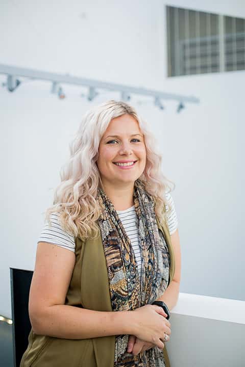 portrait photo of lecturer amy muddle standing with hands crossed and leaning on short white wall and smiling at camera with long light blonde hair and complex patterned scarf and a green sleeveless jacket