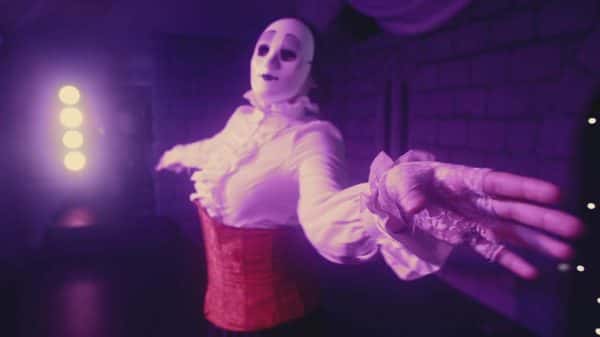 Eliza - screenshot from student film by Emma Sippitts depicts a woman in a red corset with a white frilled top, lace fingerless gloves and plastic white clown facemask in perspective shot with left hand extended towards camera and another pointing away in purple room with strong spotlight in background