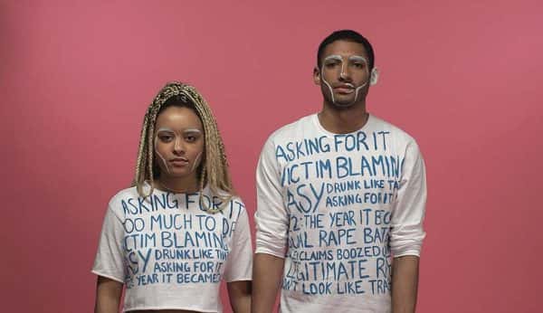 What (Rape) Culture? - screenshot taken from student film by Jade Harrison shows a man and a woman standing side by side against pink background with white shirts covered in blue writing about sexual assault against women with white paint on their faces following the chin, cheeks and brow