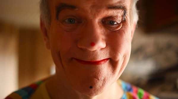 Salvo - screenshot from film by Natalie Hazelden shows close up of older man's face smiling directly into camera with red lipstick and red dot on the end of nose with raised eyebrows and grey hair