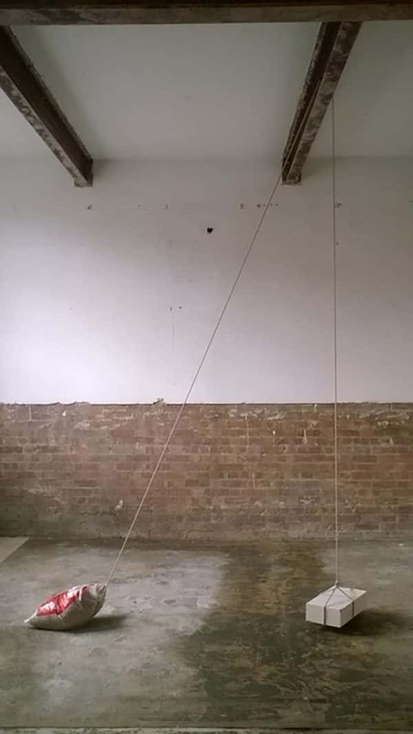  - photo shows grimy room with white upper wall and red brick lower and concrete floor with metal support beam in ceiling and a bag of sand suspending a small box in a pulley-like system