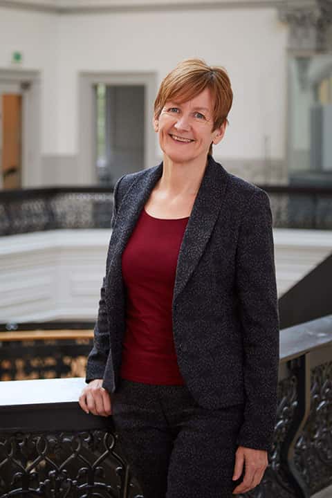 Dean of Design and Architecture at Norwich University of the Arts, Hilary Carlisle standing in Boardman House building in suit jacket & red top