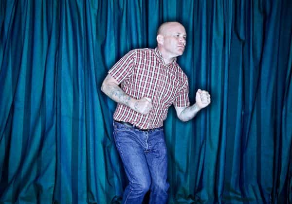 Danyelle Farrell - Man with chequered shirt in faded red and grey with blue jeans, dances with balled fists in front of a blue curtain