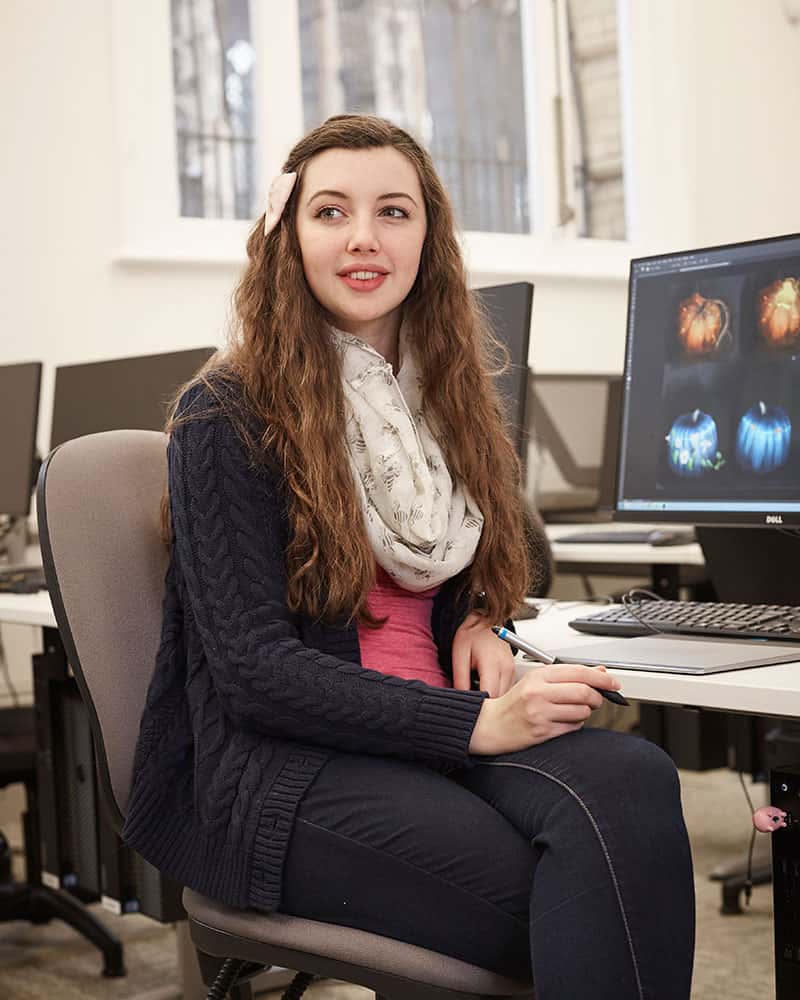 photo of alum Abigail Hookham sitting on an office chair with a digital drawing tablet pen in her hand and smiling while looking away from camera with long curly brown hair and a white loose scarf and a dark blue cardigan with several benches of computers in the background and a large window