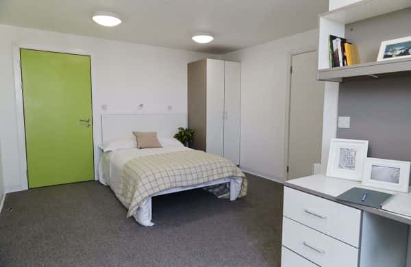 Student room - photo shows white bedroom with grey carpet and green door with bed and cushion and wardrobe in background and a desk in foreground with photos and bookshelf