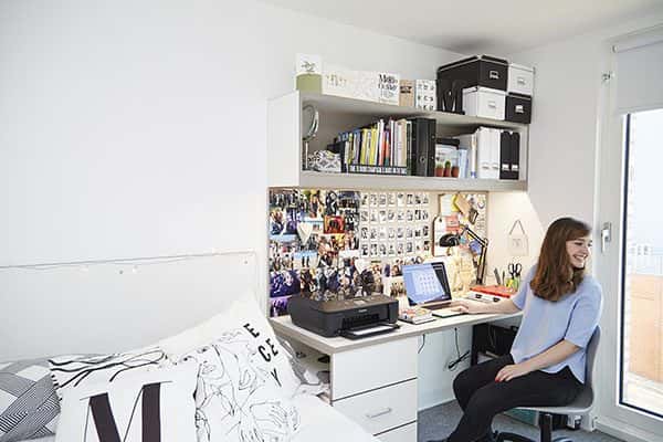  - photo shows a white bedroom with a tall window on the right hand side and a student sitting at a desk with a board of several images and paperwork and a shelf of storage items and a bed with cushions