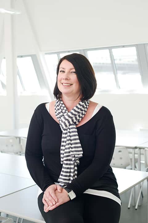 portrait photo of lecturer Gill Johnson sitting on a desk with hands on knee and smiling at camera with medium dark brown hair and a black and white striped scarf and a black long sleeved top