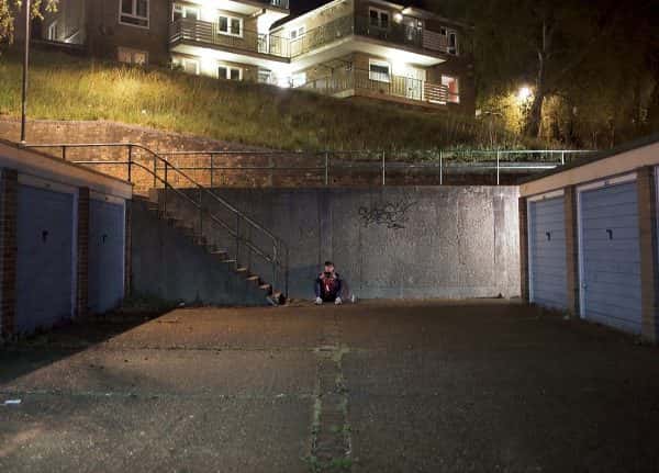 Elliott Johnson - A man in a tracksuit with a football scarf around his neck, sits next to a tin of beer on the floor of a dimly lit carpark, watching something on a mobile phone in landscape