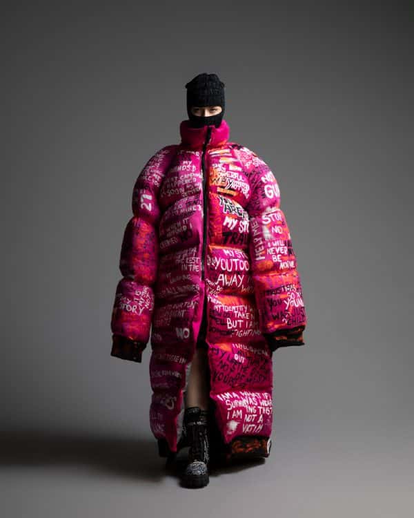 Lucy Goodall - The model is swamped in an enormous ankle-length pink puffer jacket, which is covered in bold handwritten phrases and statements.. They are wearing a black balaclava, and lace-up boots.