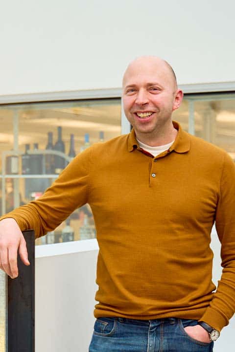 Martin Schooley, Programme Director of Communication Design, stands in an NUA building in a fulvous orange pullover with button collar, and blue jeans