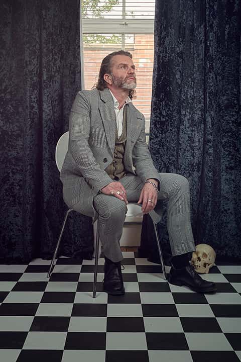 Professor Richard Sawdon–Smith — wearing a grey herringbone suit, over a waistcoat, over a white shirt which is open at the top — sits on a chair in front of a gap between two heavy dark grey curtains, letting in just a little daylight, with a skull tucked behind his foot. He looks serious and pensive.