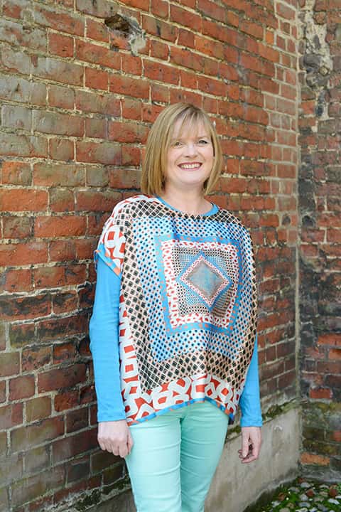 portrait photo of senior lecturer lucy blazey standing with arms by side and smiling at camera with medium blonde hair and a top with multiple square patterns reducing in size towards the centre