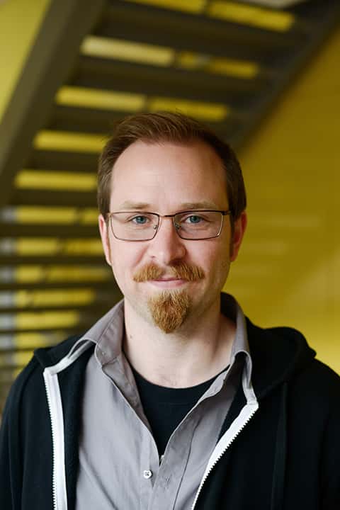 portrait photo of course leader mark wickham smiling at camera with short brown hair and light brown glasses wearing a black unzipped hoodie