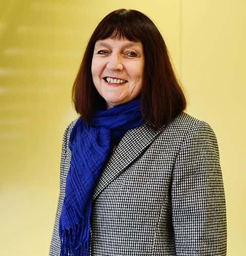 portrait photo of senior lecturer Professor Suzie Hanna standing with arms by side and smiling at camera with medium dark brown hair and a blue scarf and a buttoned black and white patterned coat