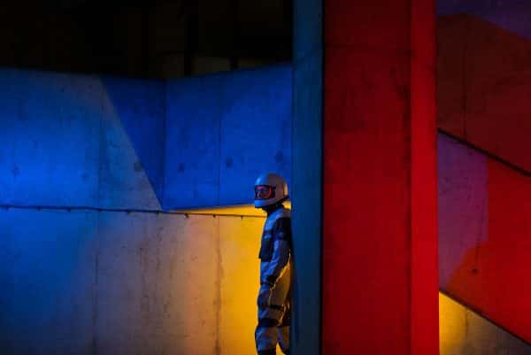 Hannah Gordon-Smith - Image depicts a man in a space suit standing in a concrete scene with red, blue and yellow lights projected onto the space