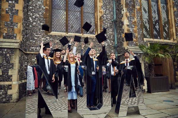  - Students throw their graduation caps into the air outside the St Andrews church building, behind a 3D installation of NUA letters made from mirrors