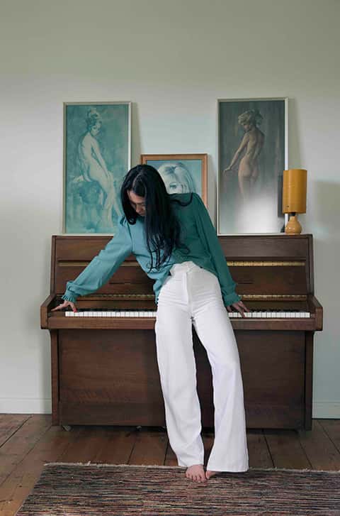 Stylist: Alex Hill. Photographer: Kerry Curl. - Photograph of a woman with long dark hair, in white trousers and a green shirt leaning against a wooden piano which has paintings propped on top
