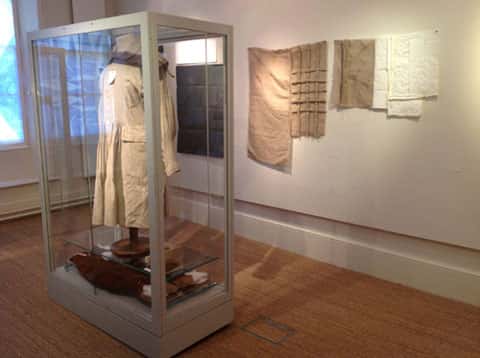 Exhibition at MEAL - Installation photo of a pale garment hanging in a glass box, behind which is beige and white fabric in rectangles hanging on the wall