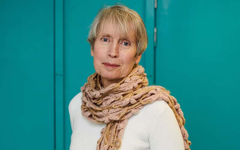 A woman with short blonde hair and a white jumper stood looking at the camera in front of a bright turquoise background