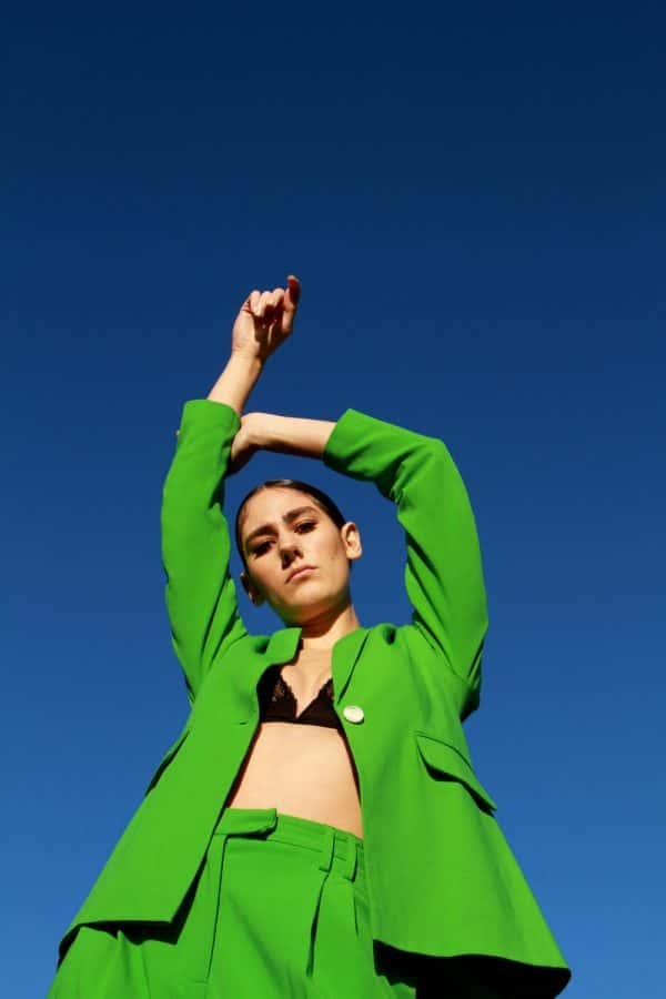 Ana Corona - Photograph of a white female model, shot from the ground looking up. She is wearing a green suit, with the jacket open and a black bra underneath. She has her hands raised above her head. Photographed and styled by BA Fashion Communication and Promotion student Ana Corona