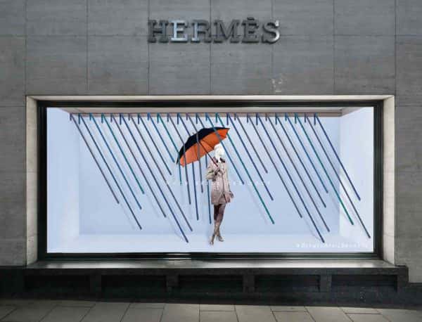 Beth Poulter - Store window visual merchandising for Hermes, featuring a sky blue backdrop, with rain falling. In the middle is a mannequin wearing a Hermes coat and holding an umbrella up. By BA Fashion Communication and Promotion student Beth Poulter