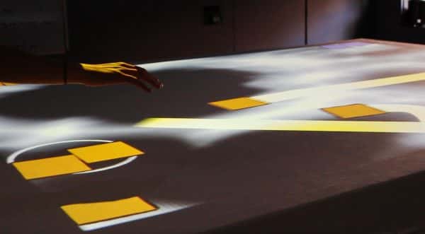  - A hand extended over a table like surface which has shapes projected on it. It appears to be a game in motion where players are 'throwing' the yellow objects back and forth across the projected image