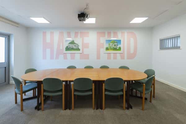  - Photograph of a conference room with a large table and chairs in the centre. On the walls is large lettering, spelling out 'Have' and 'Find', coloured pale pink. In two white picture frame centred over each of the words, is a photograph of a tree, with the word 'Strength' in one frame, and the other the word 'Peace'. The work was created by Carl Rowe, for Norwich University of the Arts' research output.