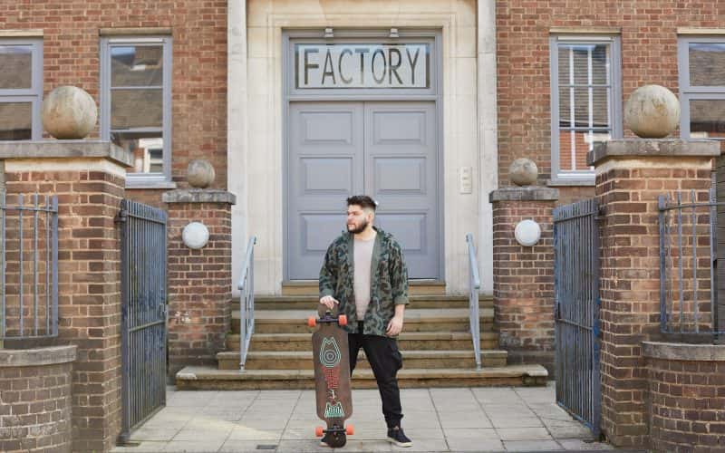BA Games Art and Design student Nikolay stands in front of the Factory Building doors in Norwich holding a long board