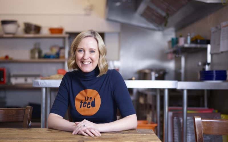 Lucy from the Feed sits wearing a Feed t-shirt looking at the camera in their kitchen