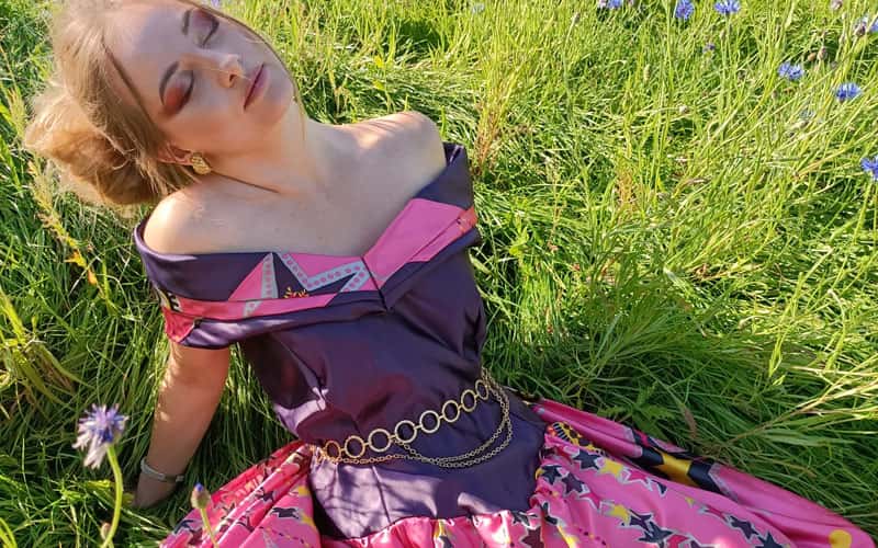 Image of a woman sat on grass, wearing a bright purple and pink off-the-shoulder dress, designed by BA Textile Design graduate Bethany Voak in collaboration with IKEA and Zandra Rhodes