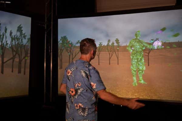 Jamie Gledhill and his interactive artwork, The Multitude - Jamie Gledhill stands in front of a screen with his hand stretched to the side, whilst wearing a blue flowery top