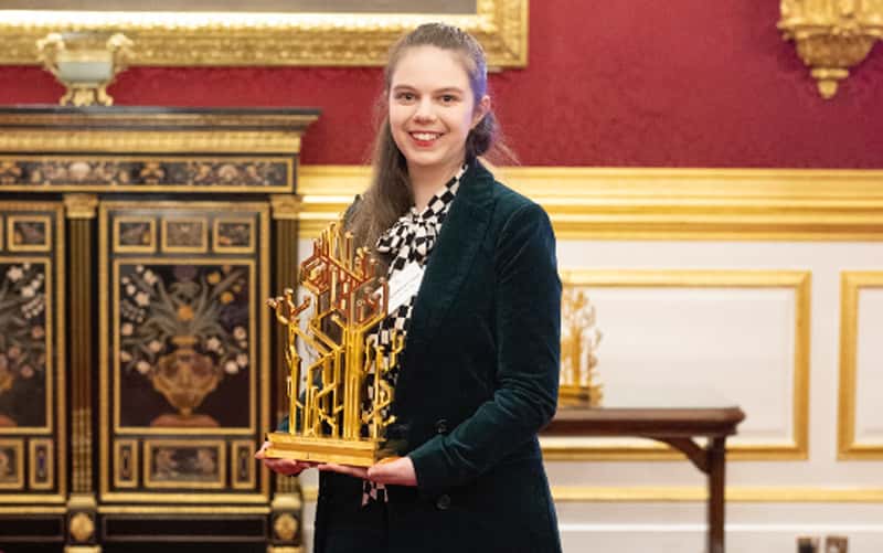 Second year BA Design for Publishing student Hannah Goldsmith, standing in the ceremony room for the QE Prize, holding the gold trophy she designed. The trophy is inspired by computer networks, and features multiple strands of metal weaving upwards and branching out into tree-like roots