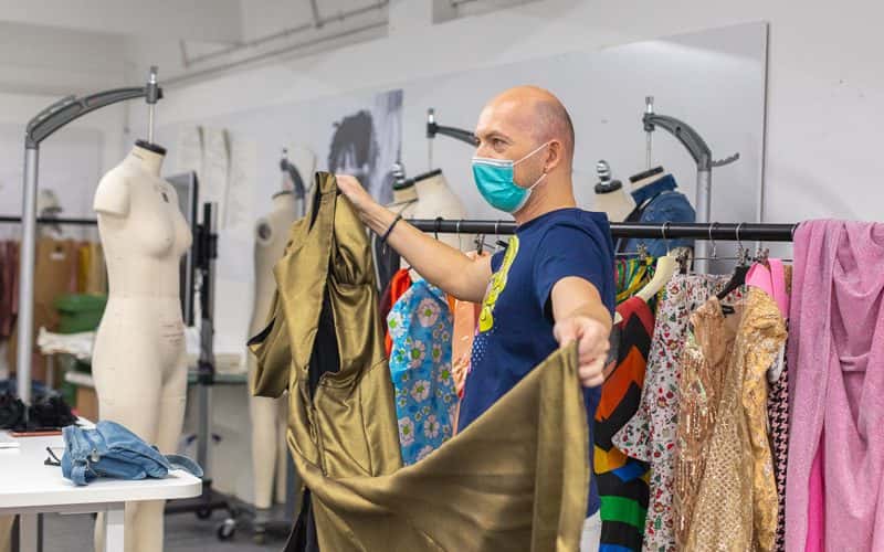 Drag queen Miss Dee Licious, in the NUA Fashion studio out of drag, displaying her own garments to students. Miss Dee Licious is pictured holding and fanning out a floor length gold dress