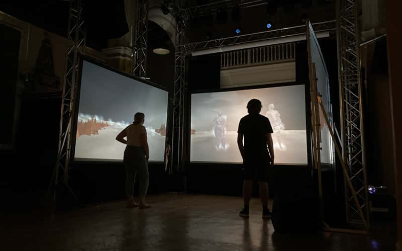 Two people standing in a dark room, in front of three screens with a projection of their bodies on