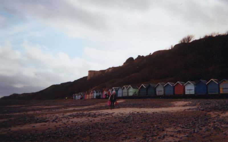 An analogue photo of someone walking beach huts by a sandy beach
