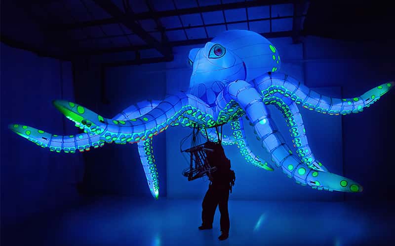 A large-scale model of an octopus glows in blue and green, while suspended by someone wearing a mechanical rig