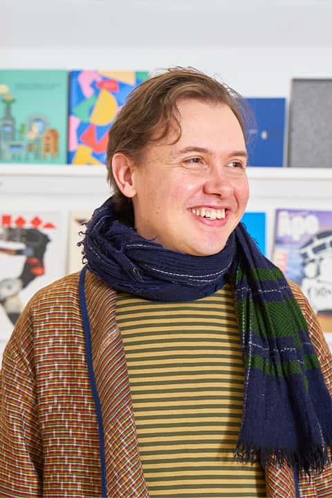 Illustration lecturer Christian Petersen smiles in front of a showcase of student work at NUA. Christian wears a navy blue scarf with green checks, yellow and grey striped shirt, and a colourful woven cardigan