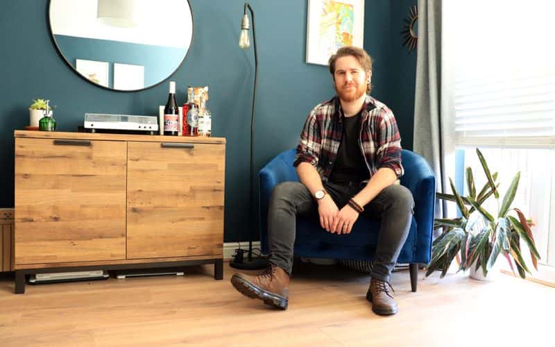 BA Graphic Design graduate Connor Edwards sat on a blue velvet chair in his home, looking at the camera. Next to him on a sideboard is a selection of his alcohol packaging designs