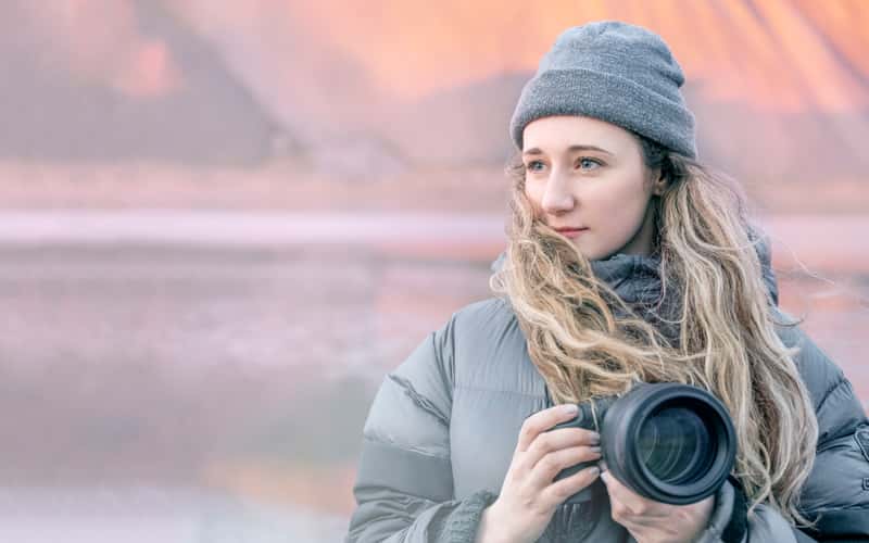 A photograph of BA Photography graduate Cat Gundry-Beck holding a DSLR camera, wearing a grey puffer jacket and grey beanie hat, looking beyond the camera