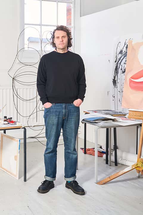 Alex Crocker standing in the Fine Art studio, wearing a black sweatshirt, blue jeans, and black doc martens