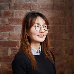 Qian Sun stands in front of a red brick wall. She has long straight hair with auburn highlights, wears round framed glasses, and a dark navy pullover over a button up shirt