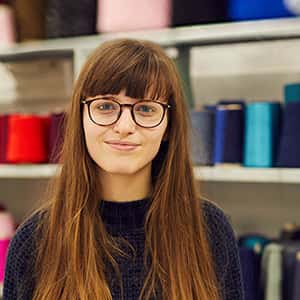 Sarah Venn stands in front of shelves of yarn and thread. Sarah wears oblong frame glasses, and has long brunette hair with a fringe.