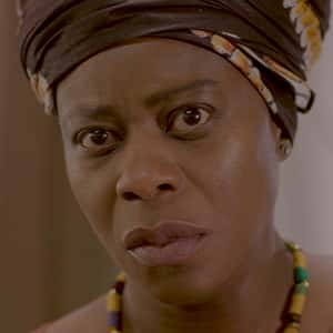 Screenshot from graduate portfolio film 'Jollof' shows a Nigerian mother, her face is full of emotion, she looks simultaneously sad, concerned, and skeptical. She wears a yellow, lilac, and green necklace of cylindrical beads, a repeating patterned top with wide neck and short sleeves, and her hair wrapped in brown cloth.,