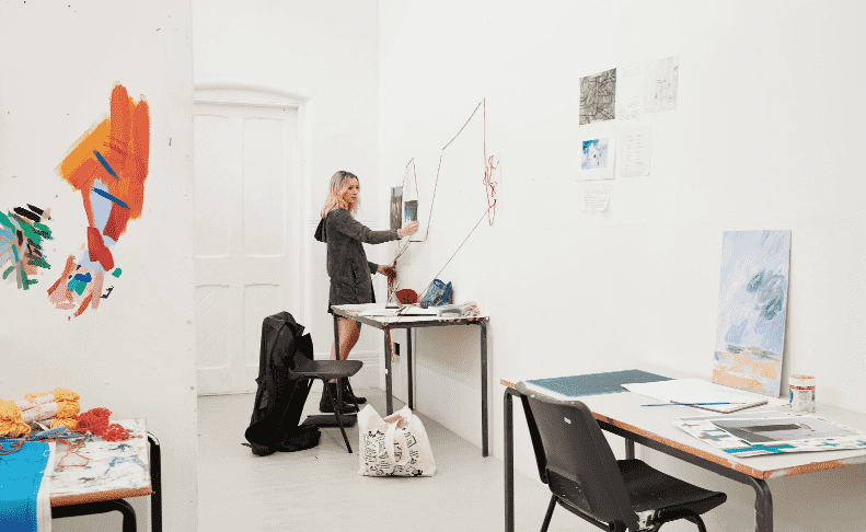 A student fixes yarn to the wall of a white studio space. Other than the white floor, walls, and fixtures, the room is dotted with colourful painting on the wall