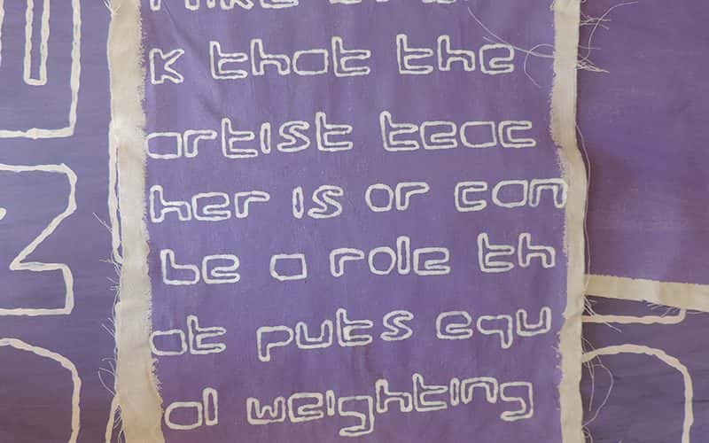 Square calico material with the words 'I like to think that the artist teacher is or can be a role that puts equal weighting on both' repeated on it overlayed on purple material