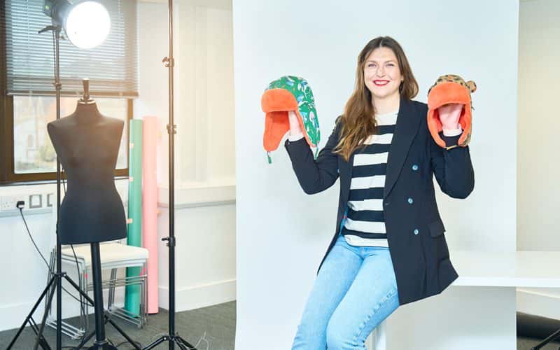 BA Fashion Marketing and Business lecturer and owner of Little Hotdog Watson, Emma Thompson, is pictured perched on the end of an empty white table, inside a fashion studio. Emma is holding both hands up, in each hand is a vibrant coloured children's hat from her own brand