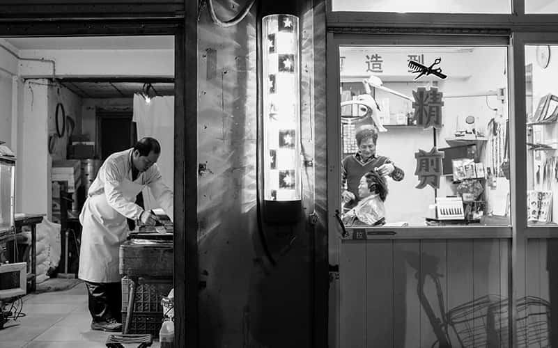 Image of two people shown working taken looking at the front of the shops. The first person is working in a hari salon cutting a clients hair and the second is preparing food.
