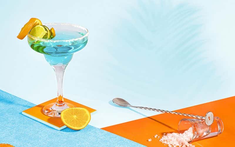 Photograph showing martini glass on a blue table with ice and blue liquid with an orange peel on the rim. Image is shot against an orange and blue backdrop.