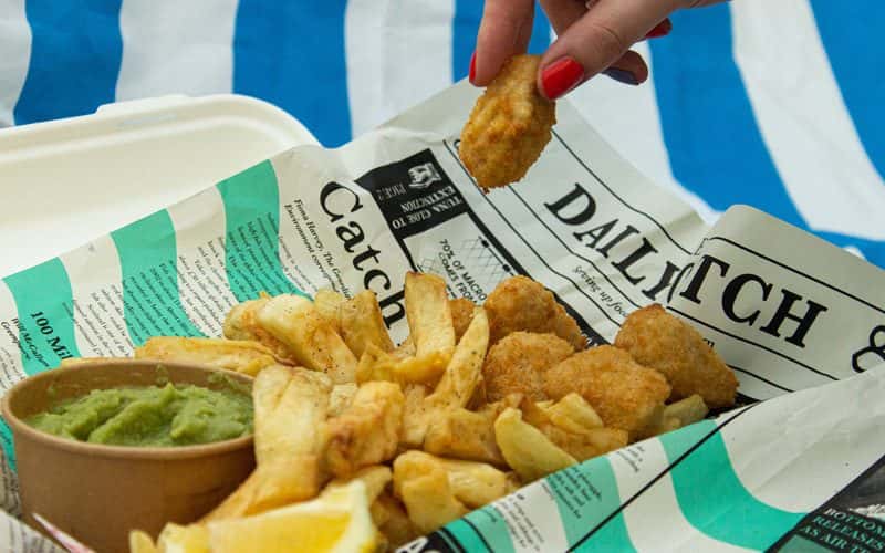 A photograph of vegan breaded scampi and chips, in traditional newspaper, with a cardboard tub of mushy peas. A hand is picking up a piece of vegan scampi. The newspaper is printed with articles on overfishing, to raise awareness of the damage caused to our oceans.
